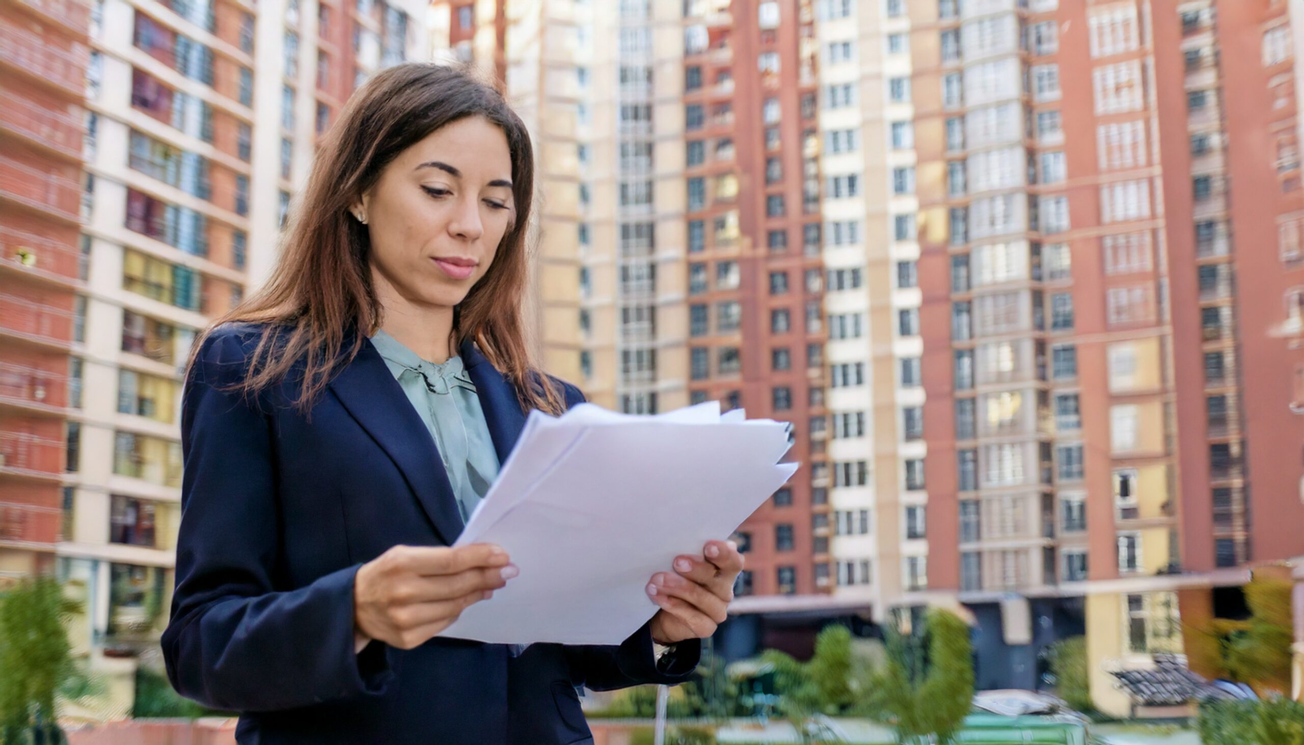 Imagem de um condomínio de fundo com síndico na frente segurando papeis de contas, despesas.